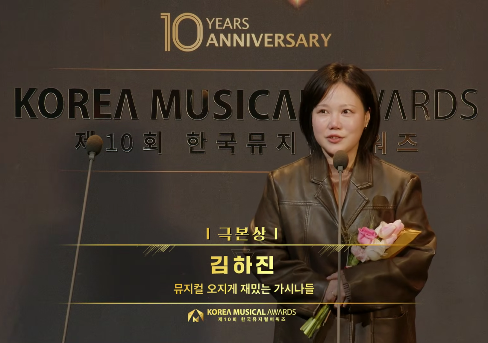 제10회 한국뮤지컬어워즈에서 극본상을 수상한 김하진 작가의 모습이다. 김하진 작가는 꽃다발을 들고 무대 위에서 수상 소감을 전하고 있다.