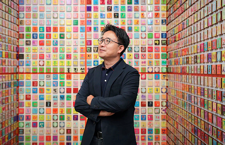 이달의 한글문화인물 ‘강익중’ 선생이 2만 개의 한글 타일로 만들어진 작품 ‘한글벽’ 앞에서 팔짱을 끼고 서 있다.