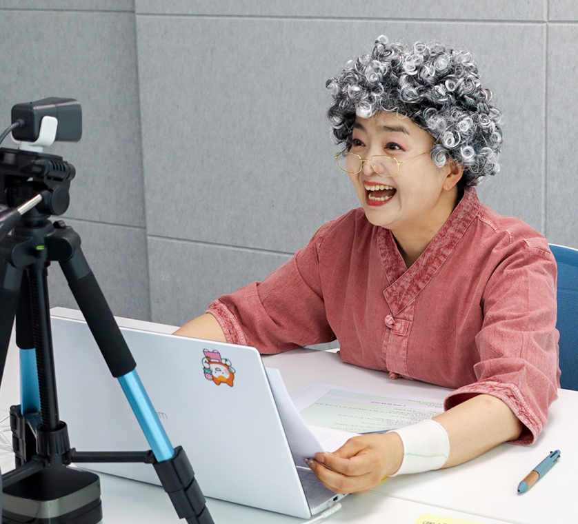 흰색 곱슬머리 가발을 착용한 ‘한글 나와라 뚝딱’ 진행자가 노트북과 카메라 앞에서 원격 한글 교육을 진행하고 있다.