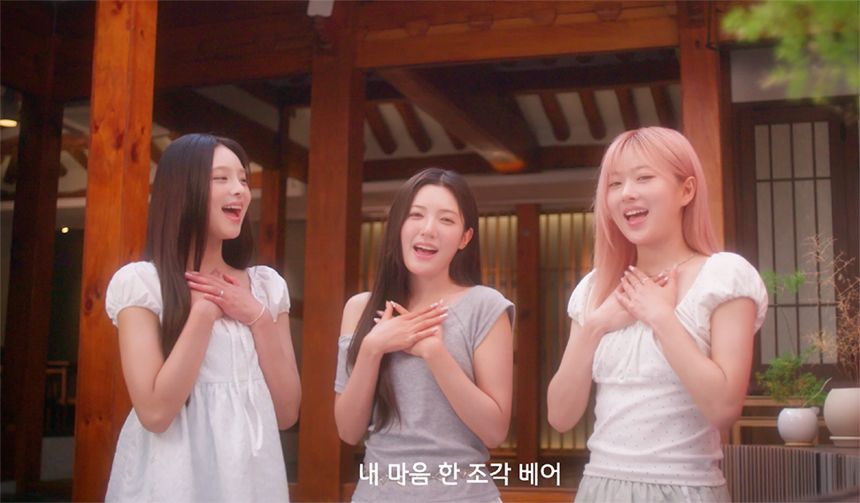 앳하트의 ‘내 마음 한 조각’ 뮤직비디오 속 장면으로, 한옥을 배경으로 세 명의 멤버가 나란히 서서 가슴에 손을 얹은 채 노래하고 있다.