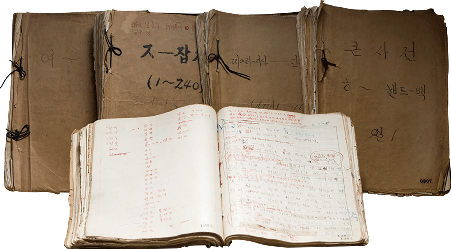 1945년 서울역 창고에서 발견된 조선말 큰사전 원고다. 낡은 갈색 표지의 원고 묶음 여러 권과 붉은색과 푸른색 글씨로 수정·교정한 흔적이 남아 있는 펼쳐진 원고 한 권이 함께 놓여 있다. 독립기념관 제공 자료다.